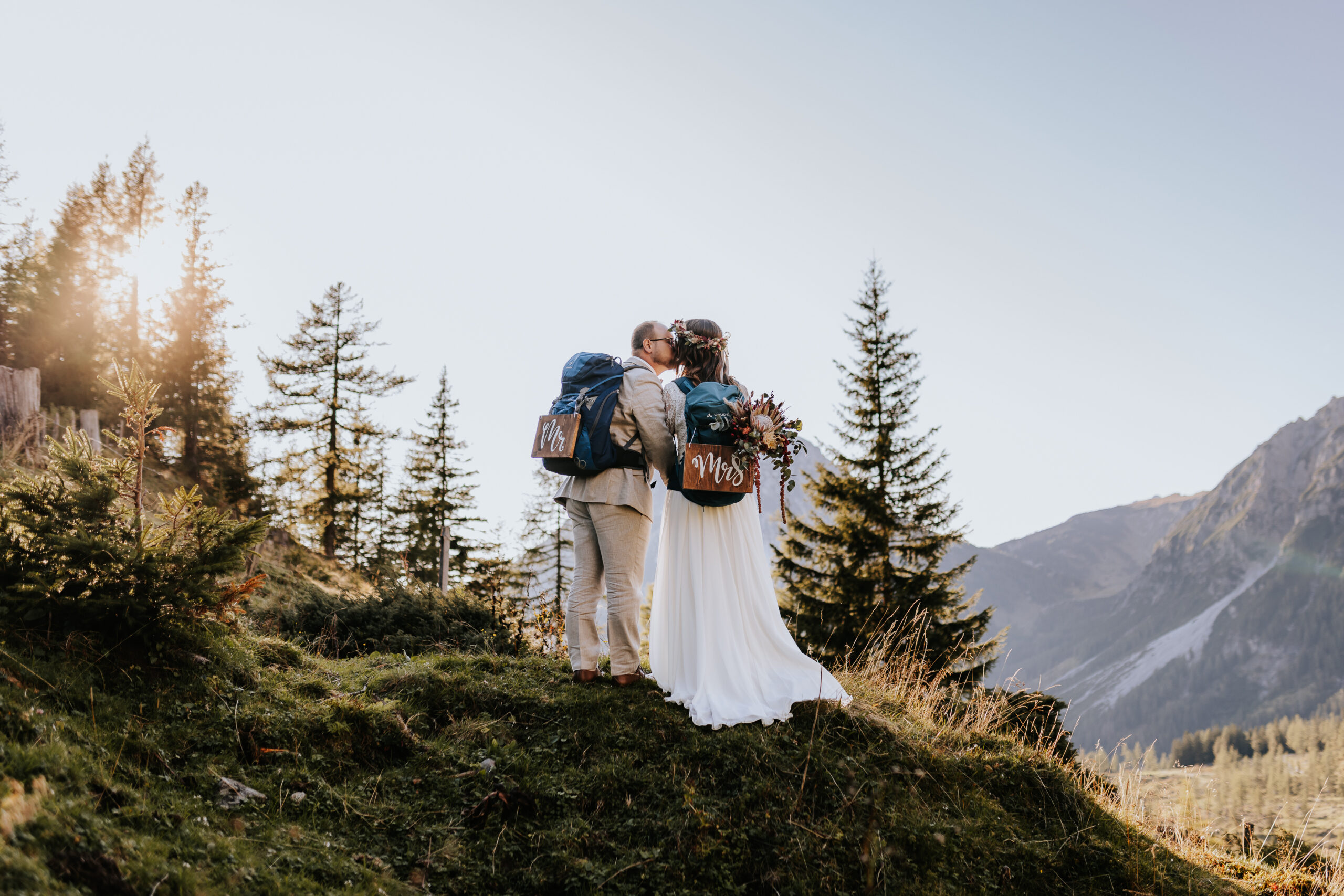 Mann in Anzug und Frau in Brautkleid küssend mit Rucksack in der Natur beim Sonnenuntergang