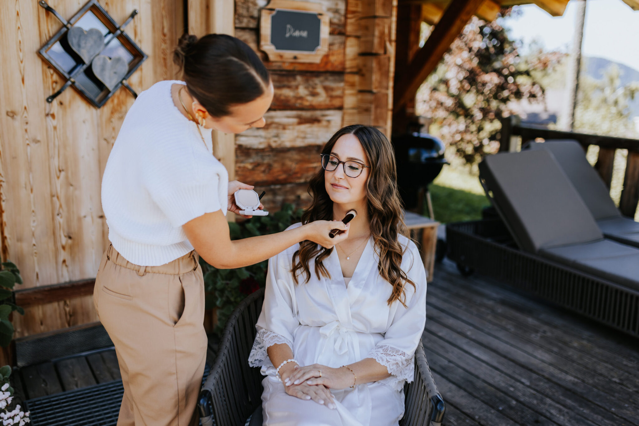 Braut im Morgenmantel mit einer Make-Up Artistin beim Schminken auf der Terrasse